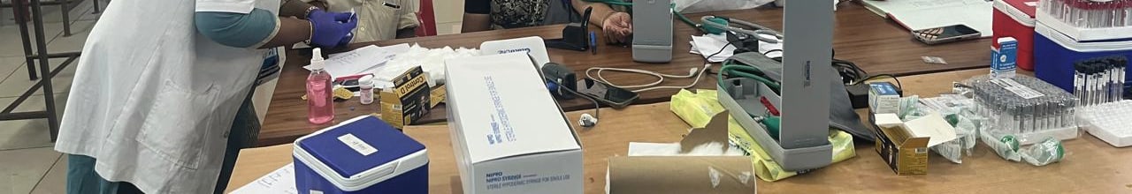 The equipment table at a community screening camp: sample tubes, cold boxes, sterile syringes and a blood-pressure monitor, with a gloved hand drawing a sample.