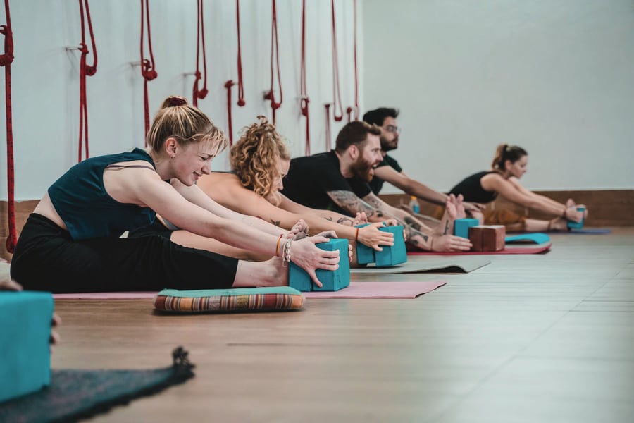 A yoga class in a studio, each person using a block to support a seated forward fold.
