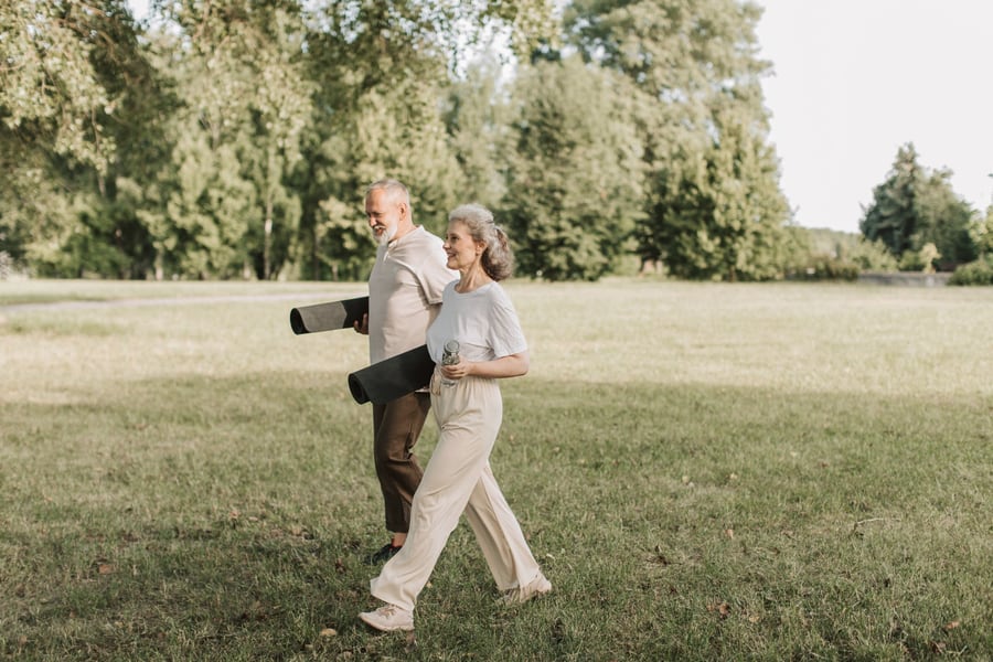 An older couple walking together across a park, carrying yoga mats.