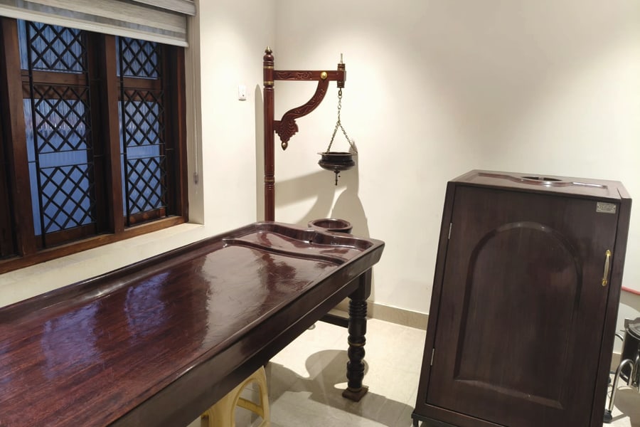 The Ayurvedic treatment room: a carved wooden droni table beneath a shirodhara stand with its hanging oil vessel, and a wooden steam chamber alongside.